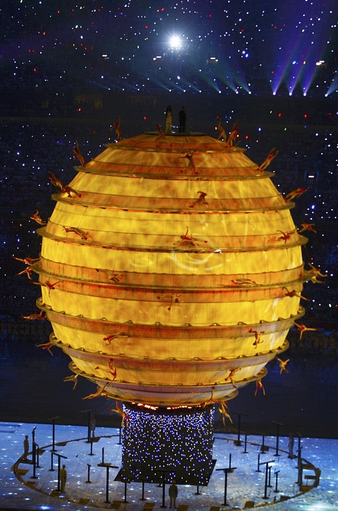 Sarah Brightman e Liu Huan fazem sueto sobre o globo que está projetando imagens e tem acrobatas correndo em volta |