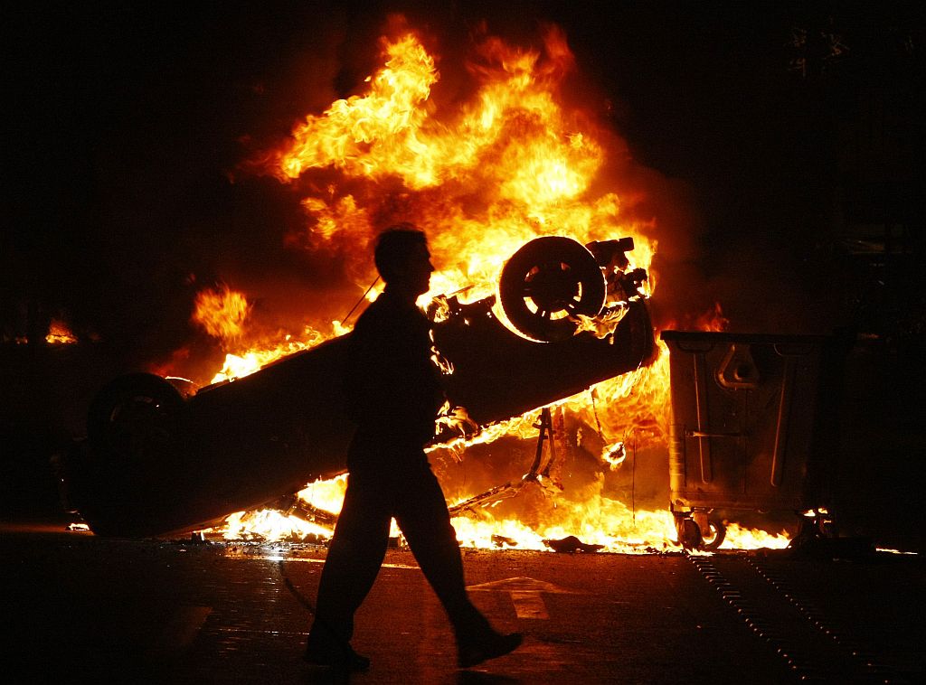 Homem caminha em área atingida pelo quebra-quebra. Ao fundo um carro foi capotado e incendiado por manifestantes | Yiorgos Karahalis / Reuters