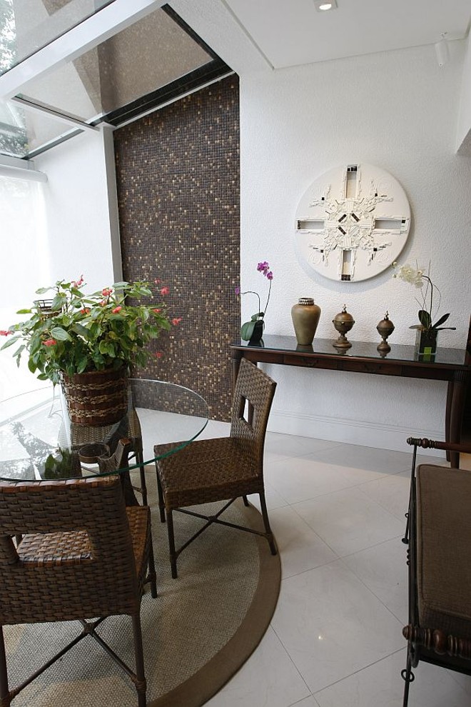 Sob a cobertura de vidro, mesa e cadeira de fibra e tapete de sisal. Na parede destaque para as pastilhas de coco e o quadro branco tipo mandala |