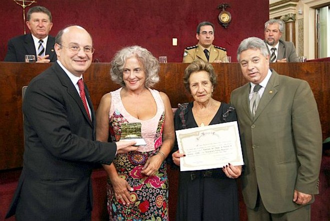 A presidente da Associação dos Amigos do Hospital de Clínicas, Maria Elisa Ferraz Paciornik (de roupa estampada), recebeu na terça-feira, em nome da entidade, o prêmio Consagração Pública oferecido pela Câmara Municipal. Quem fez a entrega foi o ex-vereador Paulo Salamuni (à dir.), que estava acompanhado de sua mãe, Hoda Salamuni, fundadora da AAHC. O reitor da UFPR, Zaki Akel Sobrinho, que compunha a mesa diretora da sessão solene, fez questão de descer para ajudar a entregar o prêmio |
