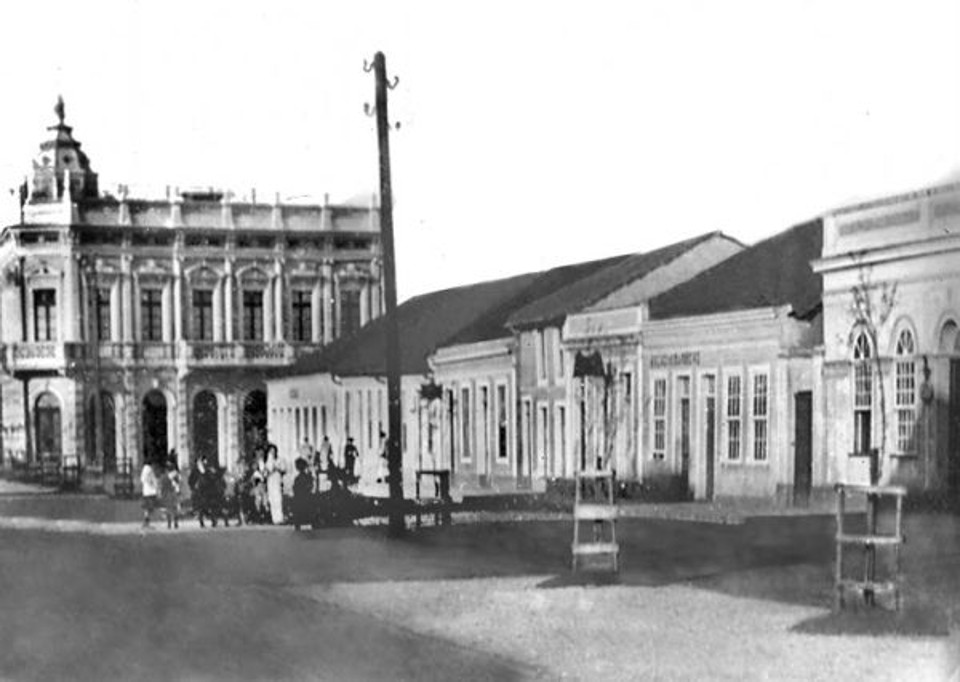 Avenida Luiz Xavier: A fotografia nos transporta ao ano de 1906, quando era prefeito de Curitiba o próprio Luiz Antônio Xavier, quando aquela via recebeu melhoramentos com a plantação de duas áleas de árvores, iluminação a arco-voltaico e calçamento dos passeios. Na imagem temos um detalhe do lado direito em direção à Travessa Oliveira Bello, onde notamos a presença do sobrado com torre em que funcionava a padaria do Müller. | 