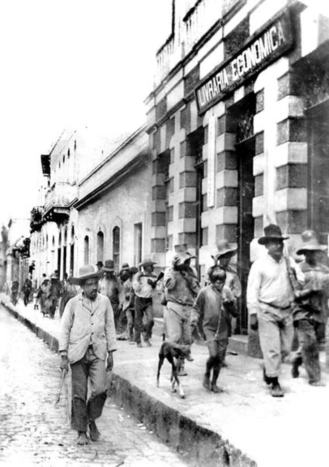 Rua XV de Novembro: Um grupo de índios, ou bugres como eram popularmente chamados, desfila pela calçada em frente à Livraria Econômica no final do século dezenove. Tais grupos de indígenas faziam presença costumeira em Curitiba, onde vinham buscar auxílio do governo. Por curiosidade, anotamos ao leitor observar a altura do meio-fio para o leito da rua. O prédio em que funcionou a Livraria Econômica, de Annibal Requião, foi a segunda sede da Gazeta do Povo, já na década de 1920. | 