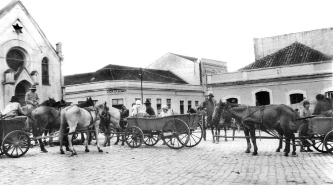 LARGO DA ORDEM Ponto de encontro dos colonos instalados na periferia do município de Curitiba. A maior frequência era da etnia polaca, que trazia seus produtos agrícolas em pequenas carroças, para ali negociarem com os estabelecimentos comerciais do local, que geralmente pertenciam a imigrantes mais antigos, os alemães. Enquanto as mulheres, depois de venderem seus produtos, faziam suas compras de fazendas e armarinhos; os homens compravam ferramentas, sal, açúcar e fumo nos armazéns e aproveitavam para dar seus taios de vudka, como apelidavam a cachaça. A foto é de 1936 | 