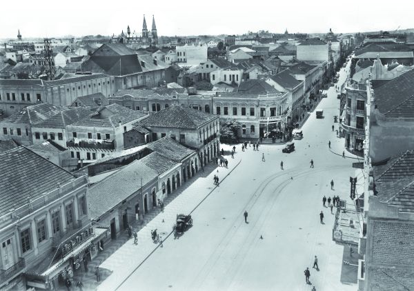 A fotografia é da Avenida Luiz Xavier e Rua Quinze de Novembro. Essa última ainda sendo mais estreita que a avenida. À esquerda, temos a torre dos Bombeiros; o telhado do Theatro Guayra se sobressai, e a Catedral aparece imponente na cidade rasa. | 