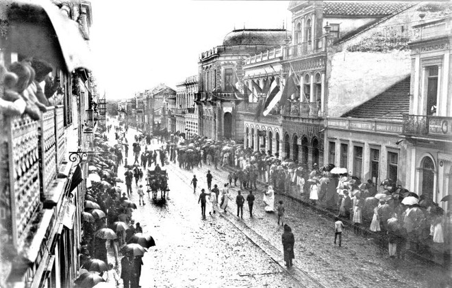 Dia 5 de março de 1912, terceiro dia de carnaval, o povo comparece abaixo de chuva para os festejos na Rua Quinze de Novembro | 