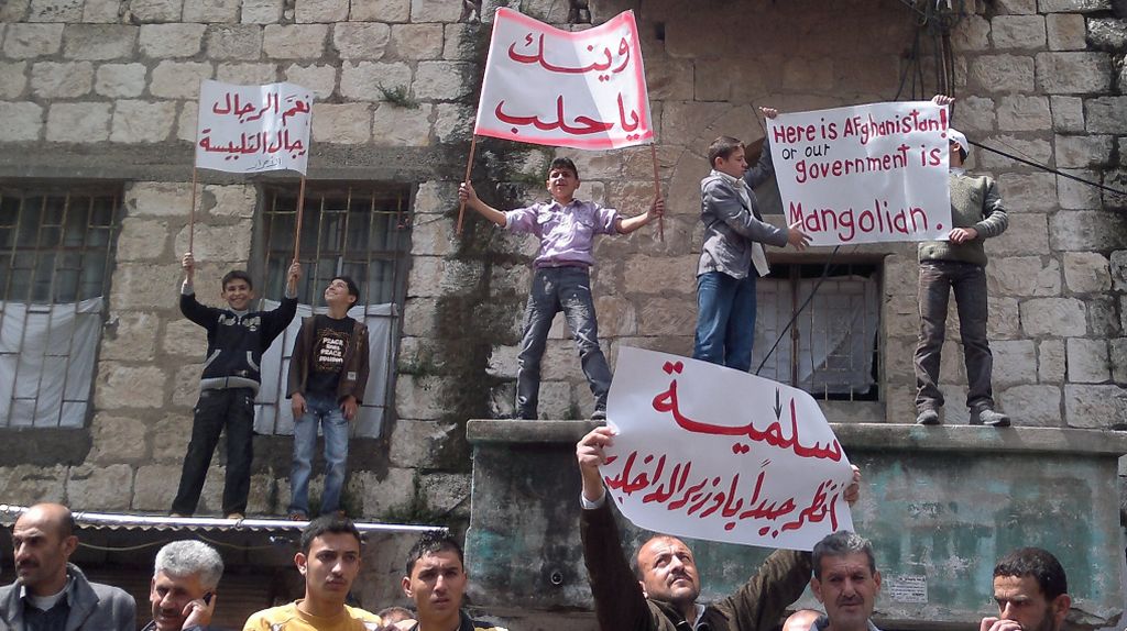 Foto tirada de telefone celular, em Banias, no nordeste da Síria, mostra protesto nas ruas da cidade: Aqui é o Afeganistão, diz uma placa, em inglês, segurada por dois meninos | AFP