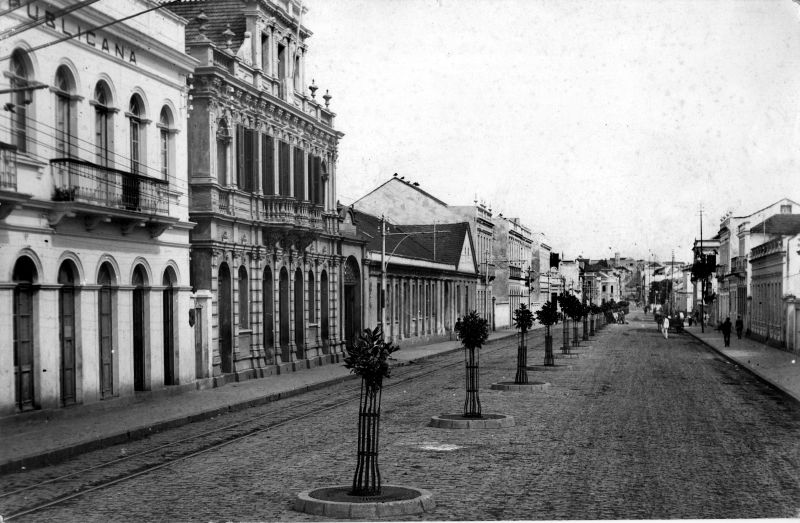 A Rua Marechal Floriano na década de 1910, quando o leito da rua foi calçado. Tendo ao centro mudas de magnólias recém-plantadas e nas laterais os passeios com pedras duquinho |