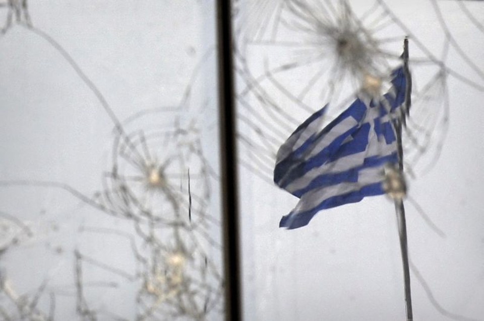 Bandeira grega vista através de vidro quebrado durante os protestos em Atenas. | AFP
