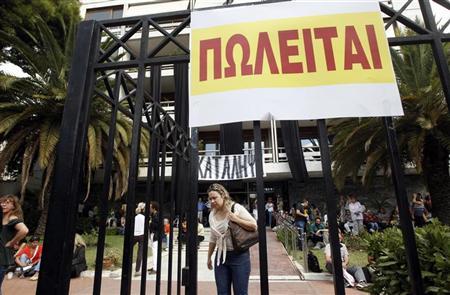 Funcionários do Ministério de Interior da Grécia sentam no pátio do prédio, bloqueando sua entrada, em Atenas. A placa lê: "À venda" | REUTERS/John Kolesidis