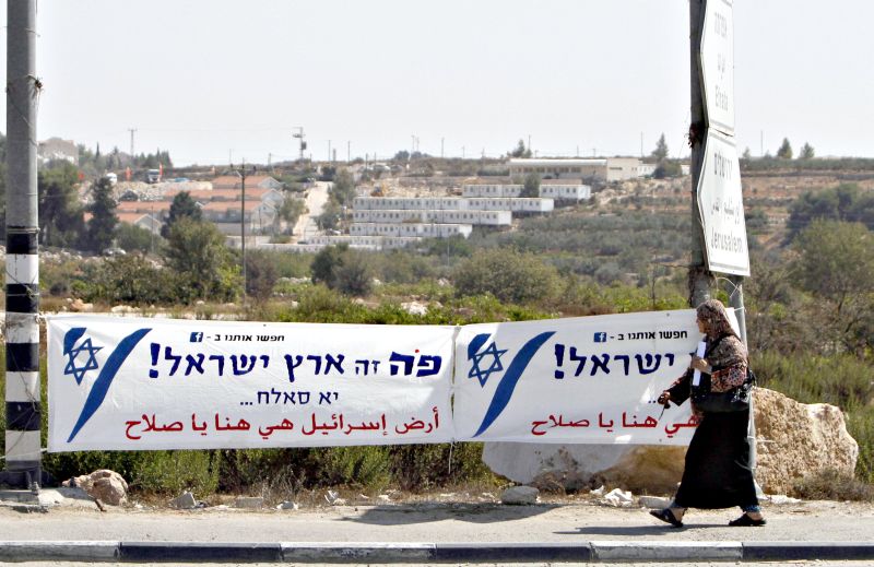 Mulher palestina passa diante de faixa em assentamento judeu com os dizeres em hebraico e árabe: Esta é a terra de Israel | Musa Al Shaer/AFP