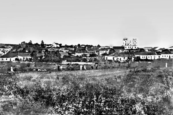Em 1870, o fotógrafo fixou essa vista parcial de Curitiba. Em primeiro plano, a capoeira onde hoje se localiza a Praça Carlos Gomes. Na margem do Rio do Ivo, vemos uma lavadeira e algumas pessoas. Ao fundo a antiga Igreja Matriz, na atual Praça Tiradentes | Arquivo CD