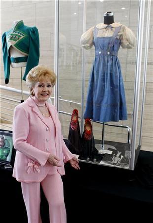 Atriz Debbie Reynolds posa ao lado do vestido azul e branco e sapatinhos vermelhos usados pela atriz Judy Garland no filme O Mágico de Oz | Fred Prouser/Reuters