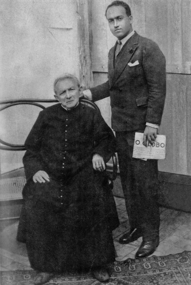 Para provar que não estava bravo depois de uma das mentiras de Benjamin, Padre Cícero tira uma foto com o sírio, que posiciona sua mão perto do cangote do sacerdote para se dizer íntimo. E segura o jornal O Globo para demonstrar intelectualidade e fazer propaganda | Reprodução