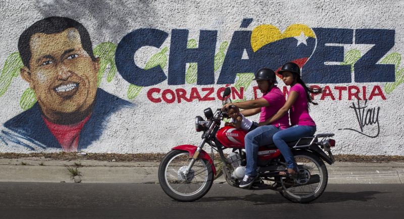 Família de moto passa por mural com a imagem do presidente Hugo Chávez, que sofre de uma infecção respiratória severa | Carlos Garcia Rawlins/Reuters