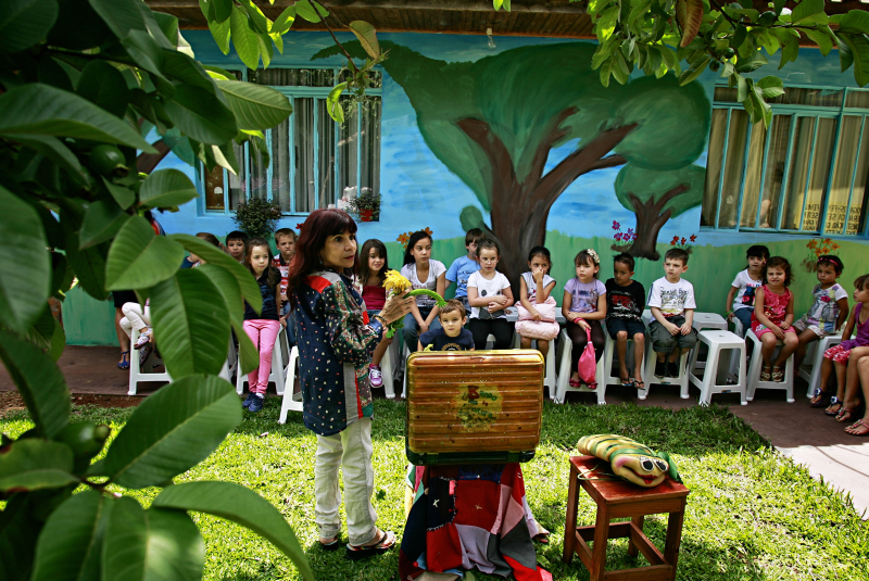 ucélia transforma a sombra de uma árvore em palco e aparece com uma valise metálica e colorida nas mãos, como se estivesse chegando de uma viagem imaginária | Josué Teixeira/Gazeta do Povo