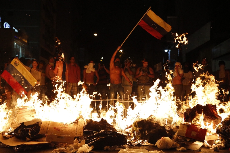 Manifestantes com bandeira venezuelana atrás de barricada em chamas, numa demonstração contra Maduro, em Caracas | Carlos Garcia Rawlins/Reuters