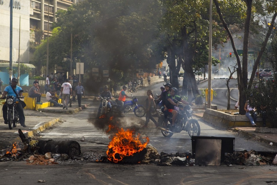 Motociclistas desviam uma barricada de fogo em Caracas. Protestos contra o governo do presidente Nicolás Maduro prosseguem na Venezuela nesta quarta-feira (19) | EFE/Miguel Gutiérrez
