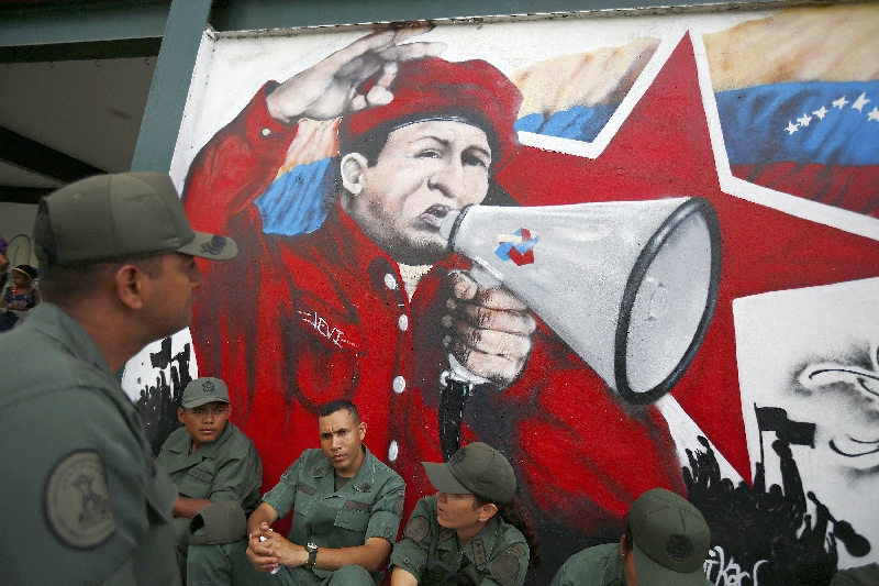 Soldados venezuelanos sentados ao pé de uma pintura do ex-presidente Chávez, que implantou medidas antidemocráticas | Jorge Silva/Reuters