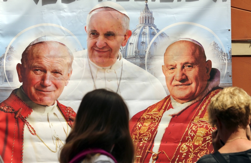 Mulher fotografa uma ilustração que reúne o papa Francisco e os dois beatos que serão santificados hoje, no Vaticano | Stefano Rellandini/Reuters