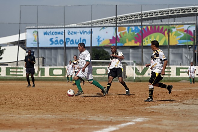 Partida de um tradicional torneio de pelada da Zona Leste de São Paulo e ao fundo as boas-vindas da Fifa para a vizinhança do Itaquerão |