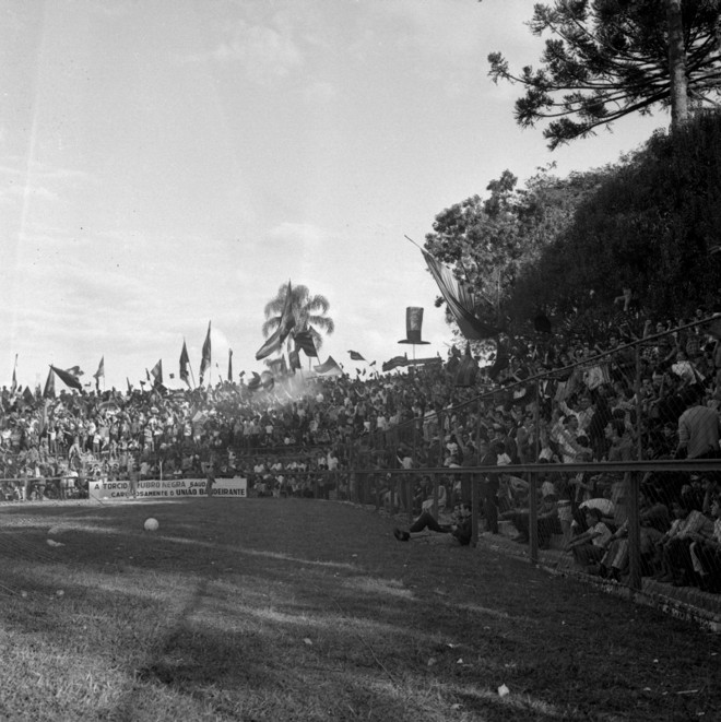 A torcida enche a arquibancada dos fundos, durante um jogo na temporada de 1968. Bandeiras, a cartola do mascote atleticano e um dos pinheiros que compunham a paisagem antiga |