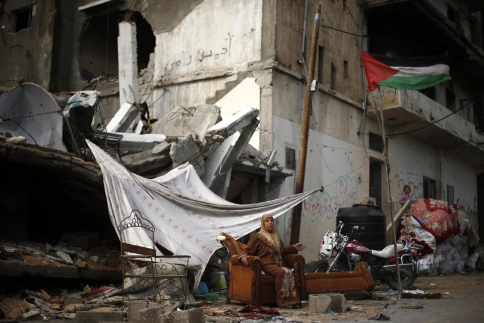 Diante da bandeira palestina, mulher senta um sofá em frente à residência, destruída nos conflitos com forças israelenses | Suhaib Salem/Reuters