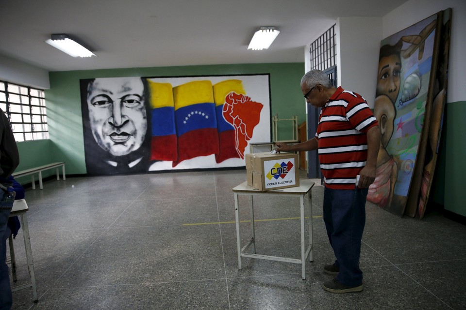 Em Caracas, um homem deposita o seu voto em uma urna perto de uma pintura do ex-presidente da Venezuela, Hugo Chávez, principal figura política do Partido Socialista. | CARLOS GARCIA RAWLINS/REUTERS