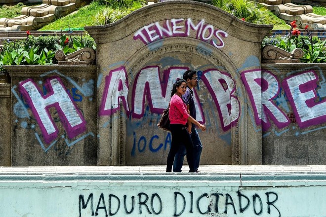 Casal caminha ao lado de muro grafitado com as inscrições “Temos fome” e “Maduro ditador”, em Caracas, na Venezuela | RONALDO SCHEMIDT/
AFP