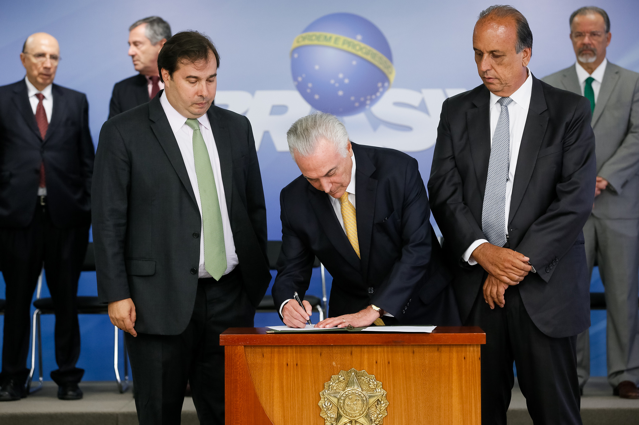 Temer entre Rodrigo Maia (esq.) e Luiz Fernando Pezão, na assinatura do decreto de intervenção federal no Rio. Ao fundo, o logotipo do governo, com o slogan “Ordem e Progresso”. | Beto Barata/PR