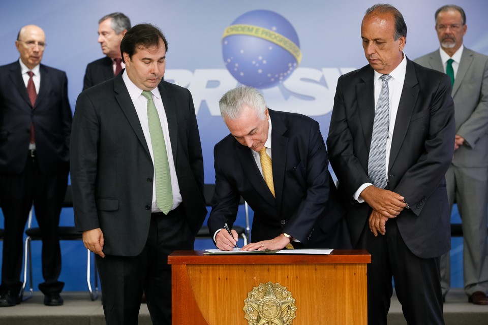 Temer ‘embute’ slogan de governo no anúncio oficial da intervenção no Rio Temer entre Rodrigo Maia (esq.) e Luiz Fernando Pezão, na assinatura do decreto de intervenção federal no Rio. Ao fundo, o logotipo do governo, com o slogan “Ordem e Progresso”. | Beto Barata/PR
