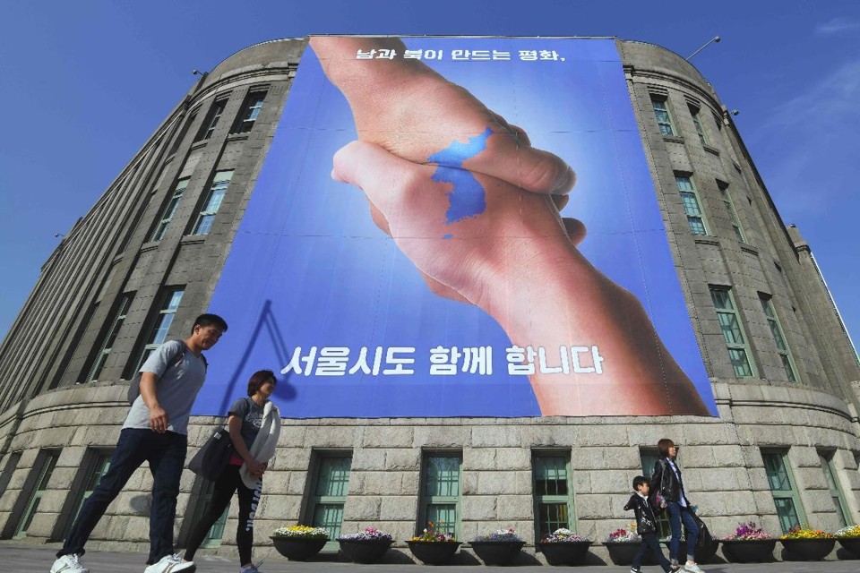 Pesdestres passam por um banner que mostra um aperto de mãos, com o formato da Península Coreana, simbolizando o desejo de um acordo entre os dois países | JUNG YEON-JE/
AFP