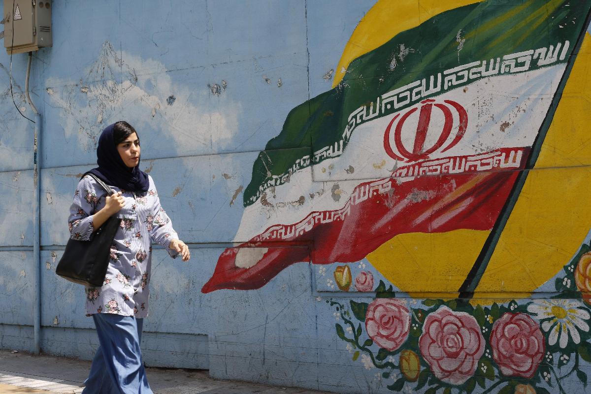 Uma mulher passa em frente a um muro onde se vê pintada a a bandeira do Irã, em Teerã | ATTA KENAREAFP