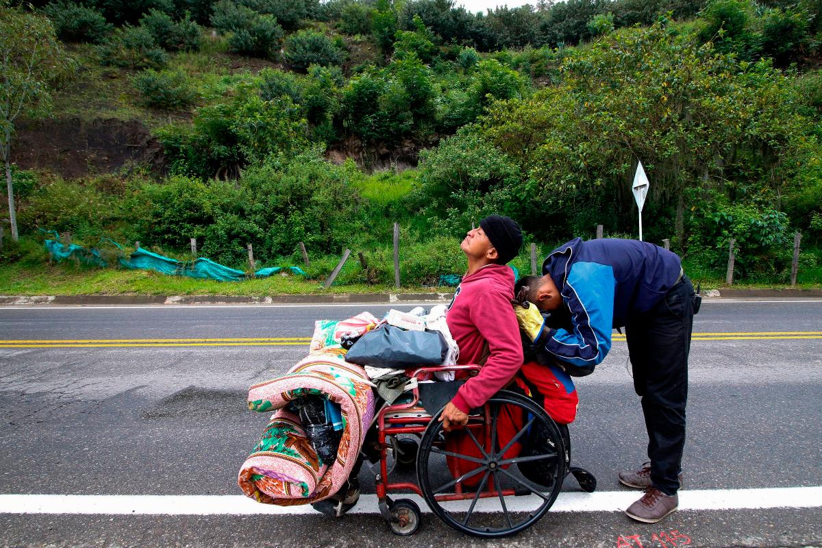 Imigrante venezuelano Jefferson Alexis e seu pai Jose Agustin Lopez, em uma cadeira de rodas, param para descansar enquanto viajam pela estrada que liga Cucuta e Pamplona, na Colômbia | SCHNEYDER MENDOZA/AFP