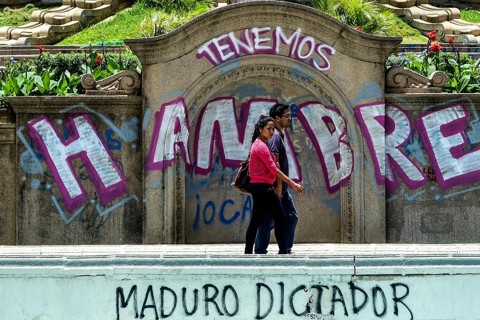 Casal passa em frente de pixação onde está escrito “temos fome” e “Maduro ditador”. em espanhol | RONALDO SCHEMIDT/AFP