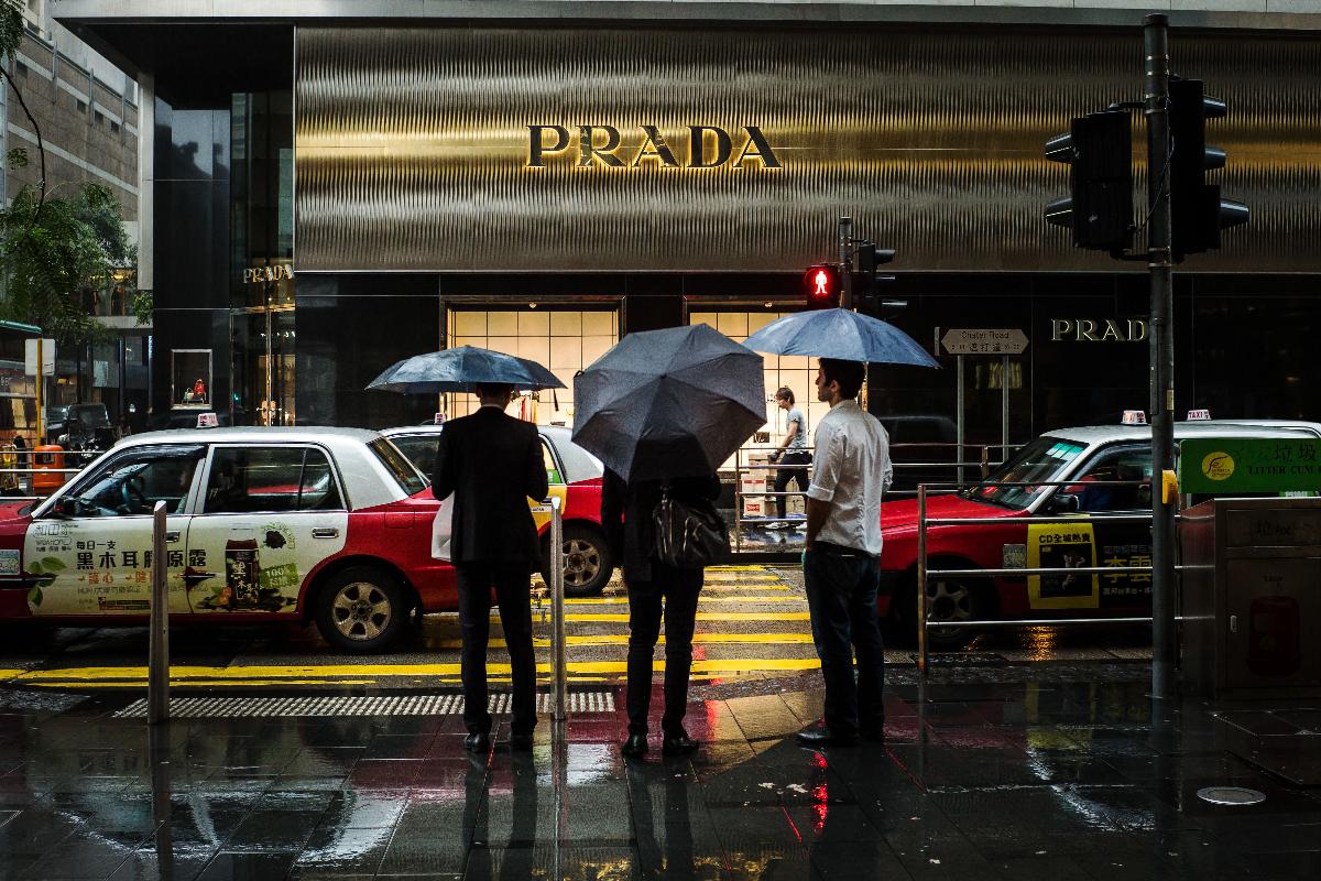 Pedestres esperam para cruzar rua em frente a uma loja de artigos de luxo da Prada, no centro de Hong Kong | Billy H.C. Kwok/Bloomberg