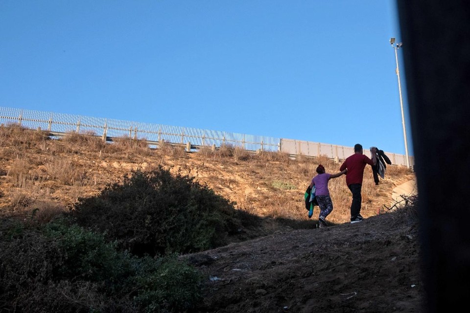 Um homem hondurenho e sua filha de dez anos caminham para se entregar à Patrulha da Fronteira, após atravessar para os EUA por um buraco embaixo da barreira de metal na fronteira, de Playas de Tijuana | GUILLERMO ARIAS / AFP