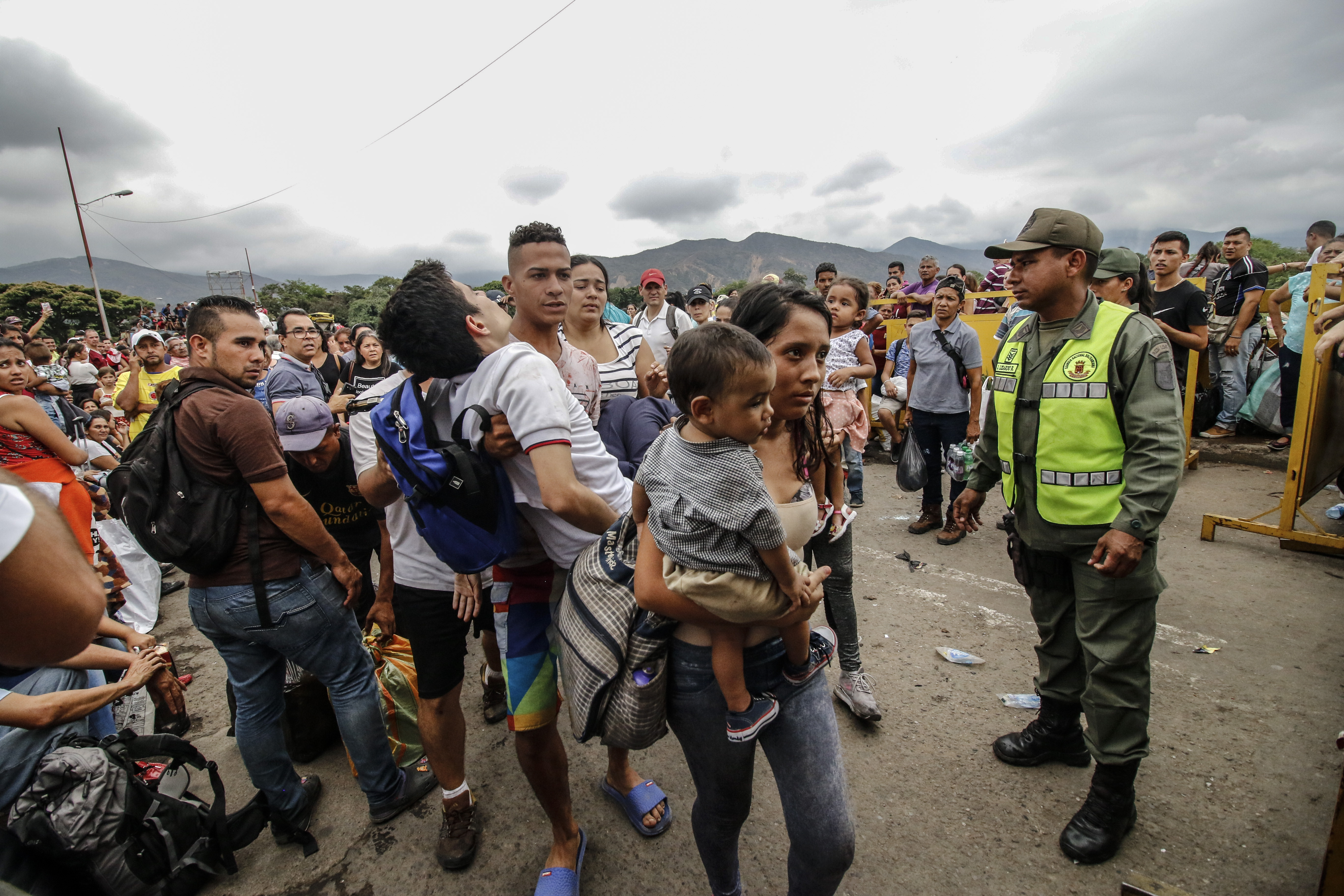 Homem leva no colo um estudante que desmaiou ao atravessar a ponte internacional Simón Bolívar, em Cúcuta, Colômbia, na fronteira com a Venezuela. Um oficial da Guarda Nacional Bolivariana os observa | Foto: Schneyder MENDOZA/AFP