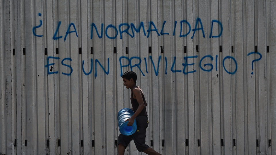 Um menino carrega um tambor com água enquanto passa por um grafite que diz "A normalidade é um privilégio?" durante uma nova queda de energia na Venezuela, na Avenida Fuerzas Armadas, em Caracas | Foto: Federico PARRA / AFP