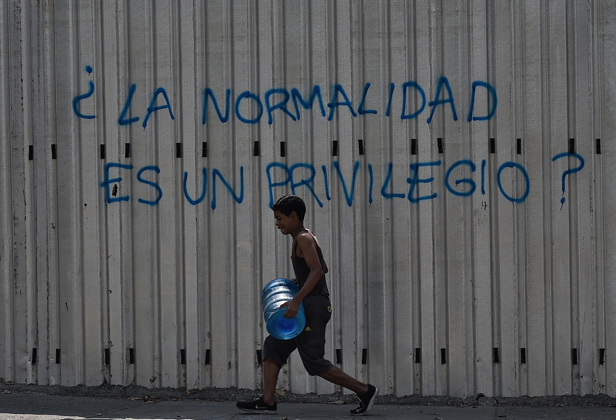 Um menino carrega um tambor com água enquanto passa por um grafite que diz "A normalidade é um privilégio?" durante uma nova queda de energia na Venezuela, na Avenida Fuerzas Armadas, em Caracas | Foto: Federico PARRA / AFP