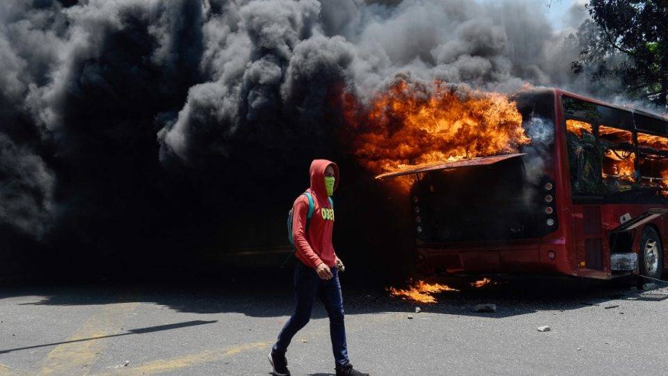Um manifestante da oposição anda perto de um ônibus em chamas durante confrontos com soldados leais ao ditador venezuelano Nicolas Maduro (Foto: Federico PARRA / AFP)
