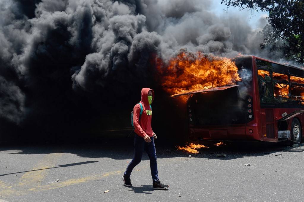 Um manifestante da oposição anda perto de um ônibus em chamas durante confrontos com soldados leais ao ditador venezuelano Nicolas Maduro (Foto: Federico PARRA / AFP)