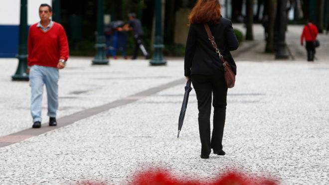 Imagem mostra uma mulher caminhando pela rua XV de Novembro, em Curitiba. Ela usa blusa fechada, bolsa na transversal e segura um guarda-chuva. Ela está caminhando, de costas para a foto. Faz frio e nublado.