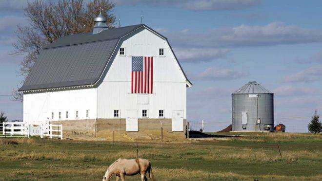 Foto mostra barracão típico de armazenagem dos EUA, de cor branca e com telhado em forma de metade de um losango. Na parede frontal uma bandeira americana. Ao lado do barracão, um silo. No primeiro plano, um cavalo pasta em relva verde.