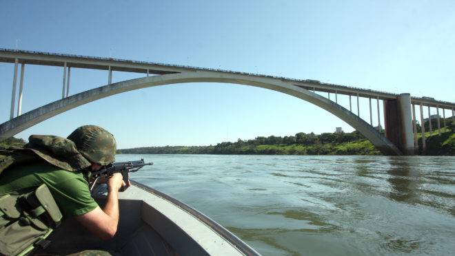 Imagem exibe soldado do Exército com carabina fazendo patrulha das margens do Rio Paraná em Foz do Iguaçu, nas proximidades da Ponte da Amizade, na fronteira com o Paraguai.