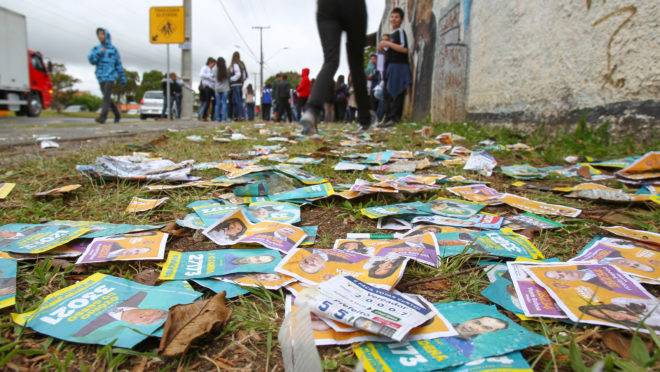 Santinhos de candidatos a vereador e prefeito de Curitiba espalhados diante de seção eleitoral.
