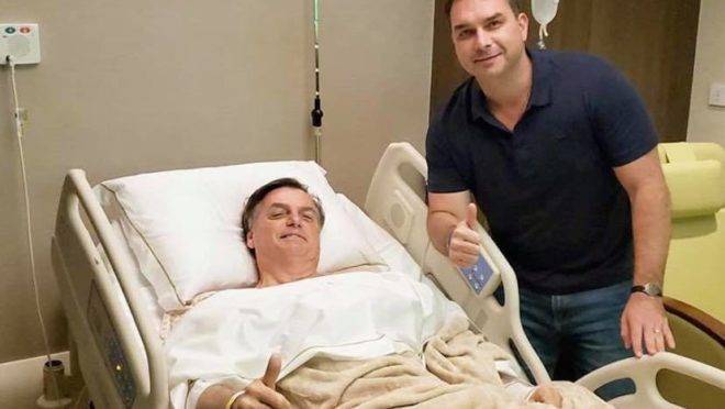 Bolsonaro na cama do hospital ao lado de Flavio Bolsonaro.
