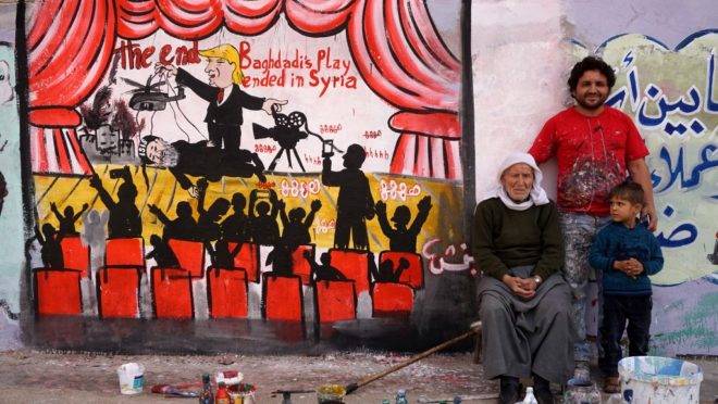 Uma foto tirada em 28 de outubro de 2019 mostra o artista sírio Aziz al-Asmar (C) posando com parentes perto de um mural que ele pintou na cidade de Binnish, no noroeste da província de Idlib, representando o presidente dos EUA, Donald Trump, dirigindo uma “peça” com a morte Abu Bakr al-Baghdadi, líder do grupo Estado Islâmico (IS), na Síria.