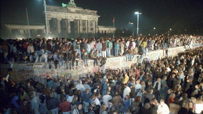Milhares de cidadãos de Berlim Oriental se reúnem no Muro de Berlim, perto do Portão de Brandenburgo (ao fundo), após a queda do Muro, 10 de novembro de 1989