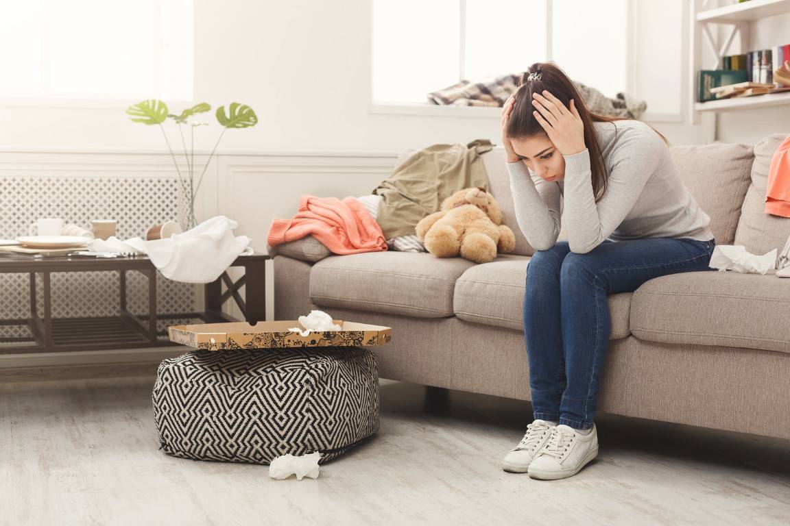Desperate helpless woman sitting on sofa in messy living room. Young girl surrounded by many stack of clothes. Disorder and mess at home, copy space Desperate helpless woman sitting on sofa in messy living room. Young girl surrounded by many stack of clothes. Disorder and mess at home, copy space