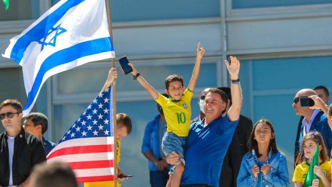 Bolsonaro acena para manifestantes a favor de seu governo em BrasÃlia com as bandeiras de Israel e EUA ao fundo.